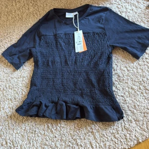 Marinblå smockad topp med volang - Säljer en marinblå topp från Gina  med smockad midja och volangkant nedtill. Toppen har korta ärmar och rund halsringning, tillverkad i mjuk bomull. Aldrig använd för den är för stor 