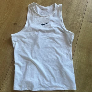 Vit Nike träningstopp i mesh tennis paddel - Säljer en vit sportig linnetopp från Nike med diskret meshstruktur och klassisk svart swoosh-logga på bröstet. Toppen är ärmlös med brottarrygg, perfekt för träning eller sportiga outfits. Materialet är lätt och andas, vilket gör den skön att bära under intensiva pass.