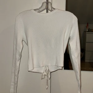 Vit ribbad croppad topp från NA-KD - Säljer en vit ribbad långärmad topp från NA-KD i storlek L. Toppen är croppad och har snygg knytning baktill. Materialet är mjukt och stretchigt, perfekt för en chill och stilren look.