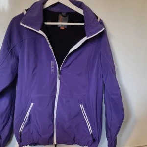 Lila vindjacka från Touch Nine - Lila jacka från Touch Nine med vit dragkedja och detaljer. Jackan har huva, två fickor med dragkedja och elastisk nederkant. Perfekt för dig som vill ha en sportig och färgstark look. Dragsko i midja.