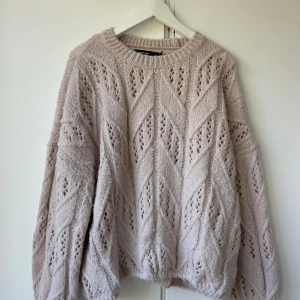 Beige stickad tröja från Vero Moda - Supermysig beige stickad tröja från Vero Moda med snyggt hålmönster och bred ribbad rund hals. Tröjan har lång ärm och loose passform, perfekt för lager-på-lager. Materialet är mjuk polyesterblandning som känns fluffigt och skönt mot huden.