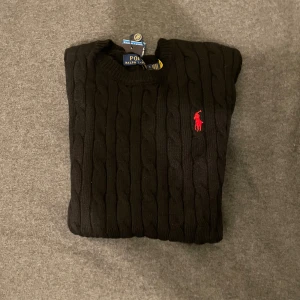 Svart kabelstickad tröja Polo Ralph Lauren - Svart kabelstickad tröja från Polo Ralph Lauren med klassisk röd logobroderi på bröstet. Rund hals och ribbade muddar vid ärmslut och nederkant. Perfekt för dig som gillar stilrena och tidlösa plagg med snygga detaljer.