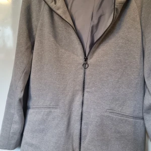 Grå hoodad jacka från ONLY, M - Grå jacka från ONLY med huva och hel dragkedja framtill. Jackan har är tillverkad i mjukt material som känns skönt mot kroppen. Enkel och clean design som passar perfekt till vardagslooken. Observera att det inte är riktiga fickor.