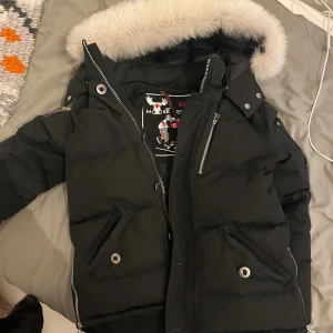 Svart dunjacka Moose Knuckles med päls - Svart dunjacka från Moose Knuckles med fluffig ljus päls runt huvan. Jackan har flera dragkedjor, snygga metallknappar och logga på ärmen. Fodrad för extra värme och perfekt för kalla vinterdagar. Klassisk pufferstil med coola detaljer.