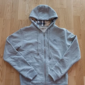 Grå Burberry hoodie med dragkedja - Snygg grå hoodie från Burberry i 100% bomull med klassiskt rutigt foder i huvan och insidan. Tröjan har huva med snörning, dragkedja framtill och två fickor. Ikonisk Burberry-logga broderad på bröstet. Passar tyvärr inte mig.