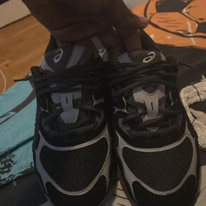 Svarta och grå Asics sneakers - Säljer ett par snygga Asics sneakers i svart och grått med coola detaljer och meshpaneler för extra andningsförmåga. Skorna har snörning och en rejäl, sportig sula som ger bra komfort. Perfekta för dig som gillar streetwear och sportig stil.