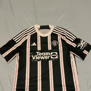 Manchester United randig fotbollströja M - Snygg Manchester United fotbollströja från Adidas med svart och vit randigt mönster och röda detaljer. Kortärmad modell med klubbmärke på bröstet och TeamViewer sponsortryck. Tillverkad i lätt och ventilerande polyester, perfekt för match eller träning. Priset kan diskuteras.