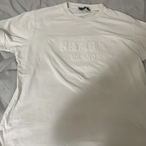 Vit t-shirt från Prada Milano - Vit t-shirt från Prada med upphöjt Prada Milano-tryck på bröstet. Klassisk rund hals och korta ärmar. Tillverkad i mjuk bomull som känns skön mot huden. Snygg och stilren design som ger en lyxig vibe till din outfit.