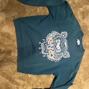 Blå sweatshirt med Kenzo tiger - Säljer en blå sweatshirt från Kenzo med det ikoniska broderade tigermotivet och loggan på bröstet. Tröjan har rund hals och långa ärmar, tillverkad i mjuk bomull. Perfekt för dig som gillar statement-plagg och coola detaljer.