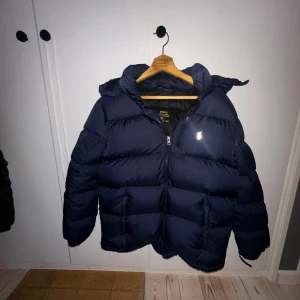 Mörkblå pufferjacka Polo Ralph Lauren  - Säljer en mörkblå pufferjacka från Polo Ralph Lauren i storlek 170 Jackan har huva, dragkedja framtill och klassisk quiltad design. Perfekt för kalla dagar och riktigt snygg streetstil. Skriv för mera bilder har två hål som jag skickar bild på privat 