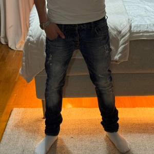 Dsquared2 jeans  - Säljer ett par snygga blå jeans från Dsquared2 med coola slitningar och tvättade detaljer. Modellen har fem fickor, klassisk knappgylf och smal passform. Jeansen har en mörkblå ton med ljusare partier och Dsquared-läderpatch bak i midjan. Skriv gärna vid fler frågor eller bilder.