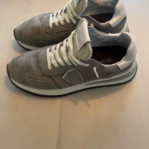 Grå Philippe Model sneakers mocka - Snygga Philippe Model sneakers i grå mocka med vita detaljer och snörning. Skorna har en rund tå, platt sula och vit logotyp på tungan. Stilren design med lyxig känsla, perfekt för dig som gillar exklusiva sneakers med italiensk touch.