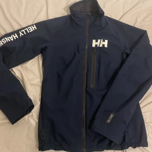 Marinblå vindjacka Helly Hansen  - Snygg marinblå vindjacka från Helly Hansen med vit logga på bröstet och ärmen. Jackan har hög krage, dragkedja framtill och en bröstficka med zip. Regular fit och tillverkad i slitstark syntet. Perfekt för blåsiga dagar och outdoor. Storlek M men sitter som S