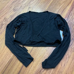 Svart croppad omlottopp med knyt - Svart croppad omlottopp med långa ärmar och knyt framtill. Tunn och mjuk, perfekt att styla över ett linne eller klänning. V-ringad design som ger en snygg siluett. Enkel och stilren, passar till många olika looks.