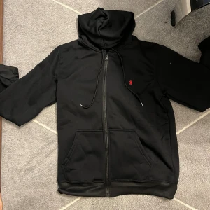 Svart hoodie från polo Ralph lauren  - Svart hoodie från Polo Ralph Lauren med dragkedja, huva och snörning. Klassisk röd logga broderad på bröstet. Mjuk och skön med känguruficka framtill. Perfekt för en chill och stilren look.