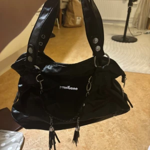 Svart handväska från Dslggao - Cool svart handväska med silverdetaljer och snygga ringar på handtagen. Väskan har dekorativa kedjor med tofsar och flera nitar som ger en edgy vibe. Perfekt storlek för att få plats med allt du behöver och passar till många olika stilar.