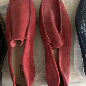 Röda mockaloafers från Oscar - Säljer ett par stilrena röda loafers från Oscar i mjuk mocka. Skorna har en klassisk rund tå och platt sula med mönstrad undersida för extra grepp. Perfekt för dig som gillar att sticka ut med färg och vill ha en bekväm och snygg sko.