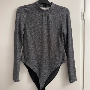 Glittrig silver body med öppen rygg - Säljer en glittrig silverfärgad body från NA-KD med lång ärm och snygg öppen rygg. Modellen har en hög halsringning och djup v-ringad rygg, samt praktiska tryckknappar i grenen. Perfekt för att sticka ut med en cool look.