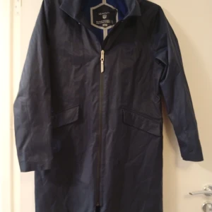 Marinblå regnjacka från Gant med huva - Snygg marinblå regnjacka från Gant med dragkedja framtill, huva och två stora fickor med lock. Jackan är lång och har en stilren look, perfekt för blöta dagar. Materialet är syntet och jackan har en klassisk passform. Str 160 medium 