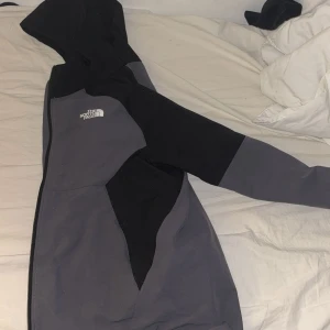 Svart och grå vindjacka från The North Face - Säljer en snygg svart och grå vindjacka från The North Face med huva och dragkedja. Jackan har en sportig look och är perfekt för blåsiga dagar. Färgblockad design och logga på bröstet ger en clean vibe. Passar dig som gillar streetwear och outdoor.