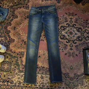 Blå slim jeans från Nudie Jeans finn 28/32 - Säljer ett par blå skinny jeans från Nudie Jeans med klassisk femficksdesign och snygga slitningar. Jeansen har mörkblå ton med ljusare tvätt och Nudie-logga på bakfickorna. Passar dig som gillar smal passform och stilren look. Midja 35cm längd 103cm (36)