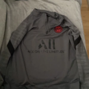 Grå långärmad PSG fotbollströja Nike - Säljer en grå långärmad Paris Saint-Germain fotbollströja från Nike med Dri-Fit material. Tröjan har svarta detaljer, PSG-logga i rött på bröstet och tryck med Accor Live Limitless samt Visit Rwanda. Perfekt för träning eller match!