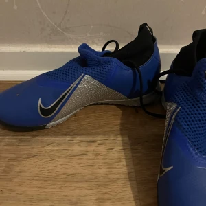 Nike Phantom Vision blå fotbollsskor - Säljer ett par Nike Phantom Vision fotbollsskor i blått med svarta och silvriga detaljer. Skorna har en modern design med meshpartier och snörning som döljs under en elastisk plös. Perfekta för dig som vill sticka ut på planen.