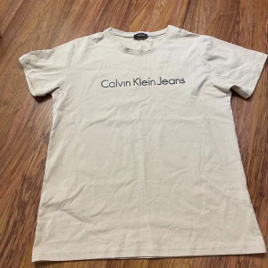 Beige Calvin Klein Jeans t-shirt - Snygg beige t-shirt från Calvin Klein Jeans med klassisk logotyp tryckt framtill. Modellen har rund hals och korta ärmar, tillverkad i mjuk bomull som känns skön mot huden. Perfekt för en clean och stilren look.