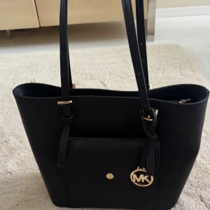 Svart axelväska från Michael Kors - Stilren svart axelväska från Michael Kors i skinn med guldiga metalldetaljer och MK-logga. Väskan har långa axelband, en praktisk ficka framtill med knappstängning och klassisk, fyrkantig form. Perfekt för dig som vill ha en snygg och rymlig väska till vardagen.