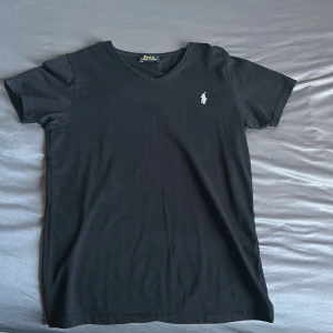 Svart v-ringad t-shirt Polo Ralph Lauren - Svart t-shirt från Polo Ralph Lauren med v-ringning och klassisk broderad logga på bröstet. T-shirten är i mjuk bomull och har en enkel, stilren design med korta ärmar. Perfekt för en clean och tidlös look.