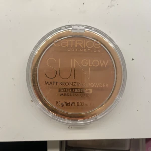 Catrice Sun Glow Matt Bronzer Medium - Matt bronzing powder från Catrice i nyansen Medium Bronze. Ger en naturlig solkysst look och är vattenresistent. Kommer i en rund plastförpackning med transparent lock och guldig kant. Perfekt för att framhäva ansiktskonturer och ge värme till huden.
