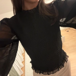 Svart blus med volang och puffärm - Supersnygg svart blus med transparenta puffärmar och smockad kropp. Blusen har volangdetaljer vid halsen och nederkanten som ger en extra touch. Köpt för något år sedan så tror ej den finns kvar på hemsidan. 