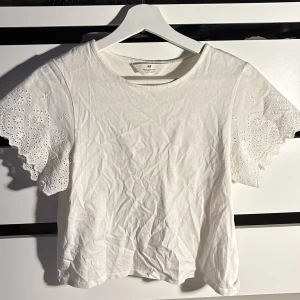 Vit blus med broderade ärmar från H&M - Säljer en söt vit blus från H&M i storlek 170. Toppen har korta ärmar med broderade blommor och hålmönster, samt en rund halsringning. Materialet är mjuk bomull. Den är skrynklig men de går att stryka 