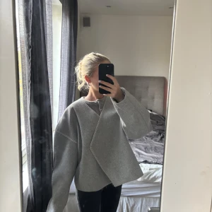 Grå omlottjacka från AWARE i storlek M - Säljer en grå omlottjacka från AWARE i storlek M. Jackan har en loose passform, lång ärm och synliga sömmar som ger en clean look. Perfekt för lager-på-lager och enkel att styla till olika outfits. Säljer pga att den är för stor.
