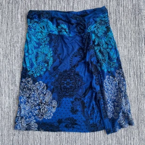 Blå mönstrad kjol från Desigual - Säljer en cool blå kjol från Desigual med detaljerat blommigt mönster i svart, turkos och vitt. Kjolen har omlott-design och en liten slits framtill. Materialet är mjukt och stretchigt, perfekt för dig som gillar färgstarka plagg med unik stil. Mått Midjan: ca 38 cm Total längd: ca 58 cm  100% viskos  Kommer från ett djur och rökfritt hem.