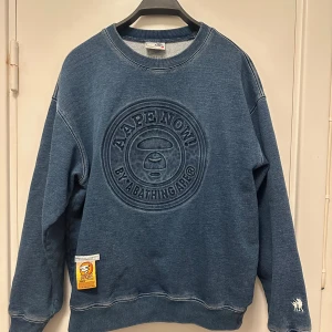 AAPE BY A BATHING APE - Säljer en blå sweatshirt från AAPE BY A BATHING APE med stort präglat logotyptryck på bröstet och broderad detalj på ärmen. Tröjan har ribbade muddar och en patch nertill. Perfekt för dig som gillar streetwear och snygga detaljer.