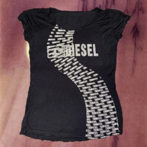 Svart topp med tryck från Diesel - Cool svart topp från Diesel med korta ärmar och ett stort, glittrigt logotryck framtill. Mönstret består av flera små Diesel-loggor i vitt som bildar en vågig form över hela framsidan. Perfekt för dig som gillar streetstyle och vill sticka ut.