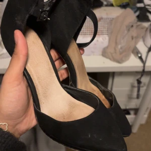 Svarta pumps med spetsig tå - Snygga svarta pumps  med spetsig tå och smal klack. Skorna har en elegant rem med spänne runt vristen och är tillverkade i mocka-liknande material. Perfekta för dig som vill ha en klassisk och stilren look.