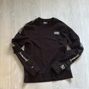 Svart EA7 sweatshirt med logga - Svart sweatshirt från EA7 Emporio Armani med tryckta loggor i vitt och gult längs båda ärmarna och en liten EA7-logga på bröstet. Tröjan har rund halsringning och långa ärmar. Perfekt för dig som gillar streetwear och snygga detaljer.