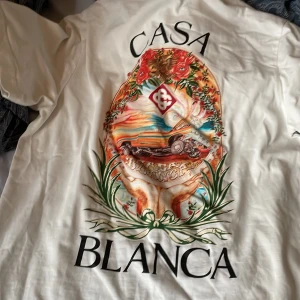 Vit Casa Blanca skjorta med tryck - Säljer en vit skjorta från Casa Blanca med stort färgglatt tryck på både fram- och baksidan. Trycket har motiv med bil, blommor och konstnärliga detaljer. Skjortan har klassisk passform och är tillverkad i mjuk bomull.