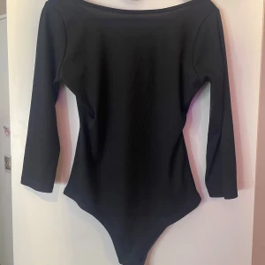 Svart ribbad långärmad bodysuit - Svart bodysuit med ribbad struktur och lång ärm. Snygg, djup urringning med dekorativa band framtill som ger en cool detalj. Stretchigt material som sitter tight och framhäver figuren. Perfekt att styla med jeans eller kjol för en trendig look. Aldrig använd 