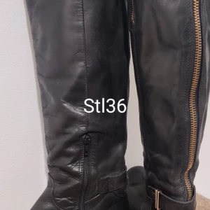 Svarta skinnstövlar med dragkedja stl 36 - Snygga svarta stövlar i skinn med grov dragkedja i guld längs sidan och dekorativt spänne vid ankeln. Platt sula och rund tå ger en cool och avslappnad vibe. Perfekta till jeans eller kjol för en edgy look.