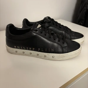 Svarta Philipp Plein sneakers i skinn - Snygga svarta sneakers från Philipp Plein i äkta skinn med metallplatta på plösen och logga på hälen. Skorna har snörning, vit platt sula med nitar och rund tå. Perfekt för dig som gillar exklusiv streetstyle och detaljer som sticker ut. Skorna är även Limited edition.