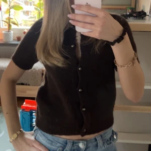 Brun stickad kortärmad tröja - Säljer en brun stickad tröja med korta ärmar och knappar framtill. Modellen är croppad och har en klassisk rund halsringning. Perfekt att styla med jeans eller kjol. Ej stickig 