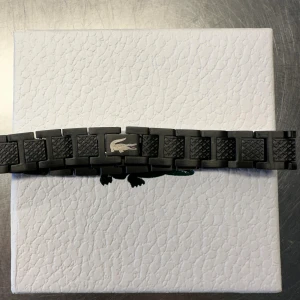 Lacoste armbandsur  - Snyggt armbandsur från Lacoste med länkarmband i metall. Finns i svart, silver och guld med den klassiska krokodilloggan på spännet. Länkarna har en cool textur som ger en modern vibe. Perfekt accessoar för dig som gillar stilrena detaljer.