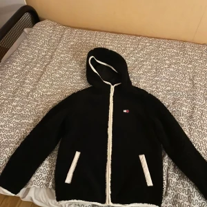 Svart teddyjacka från Tommy Jeans - Svart teddyjacka från Tommy Jeans med huva och vita detaljer längs dragkedja och fickor. Jackan har broderad logga på bröstet och är tillverkad i mjukt teddy-material. Perfekt för kalla dagar och riktigt snygg streetstil. Priset kan diskuteras 