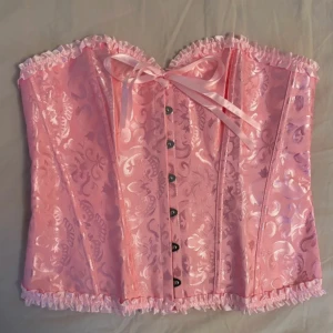 Rosa korsett med rosett och spetsdetaljer - Supersöt rosa korsett med glansigt mönster, volangkant och rosett framtill. Toppen har snörning i ryggen och knäppning fram. Materialet är polyester med elastan för stretch. Perfekt för dig som vill sticka ut med en feminin och trendig look.