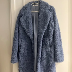 Ljusblå teddyjacka från Gina Tricot - Supermysig ljusblå teddyjacka från Gina Tricot med fluffig yta och stora slag. Jackan har en oversized look och är perfekt för att hålla sig varm och snygg under kyliga dagar. Fodrad insida och två fickor framtill.