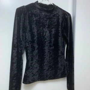 Svart sammetstopp med lång ärm - Säljer en svart långärmad topp i mjuk sammet med hög hals. Toppen har en figurnära passform och en stilren look som passar perfekt till jeans eller kjol. Materialet ger en lyxig känsla och den svarta färgen gör den lätt att matcha.