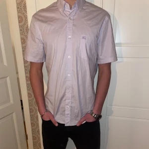 Burberry skjorta - Snygg ljuslila skjorta från Burberry med korta ärmar och klassisk krage med rutigt mönster på insidan. Skjortan har knappar framtill och en diskret logga på bröstet. Skjortan är i bra skick (pris kan diskuteras).